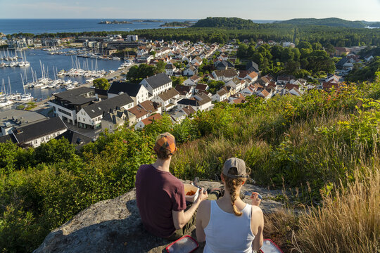 View of a couple sitting on a rocky hill overlooking the town of Mandal with its marina, white wooden houses, and lush greenery in Mandal, Agder, Norway.