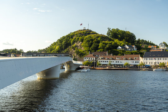 View of the modern white Mandal Bridge spanning the river with historic white wooden houses and a green hill in the background in Mandal, Agder, Norway.