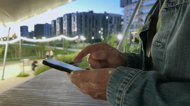 Smartphone in hand, a man scrolls news and reads updates while standing beneath a canopy in an evening city park. Modern residential towers rise in the background as he browses his feed.