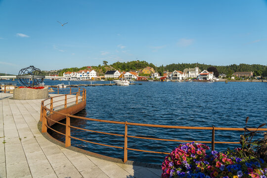 View of the harbor in Mandal with colorful buildings, boats, a modern sculpture, and vibrant flowers in the foreground under a clear blue sky in Mandal, Agder, Norway.