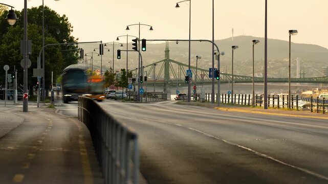 Budapest, Hungary - August 1, 2026: A static wide-angle time lapse capturing the warm golden hour atmosphere along a Budapest riverside road, with moving commuter traffic, street lights, and the histo
