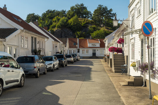View of a quiet street in the town of Mandal, featuring traditional white wooden houses with red tiled roofs, parked cars, and blooming flower boxes in Mandal, Agder, Norway.