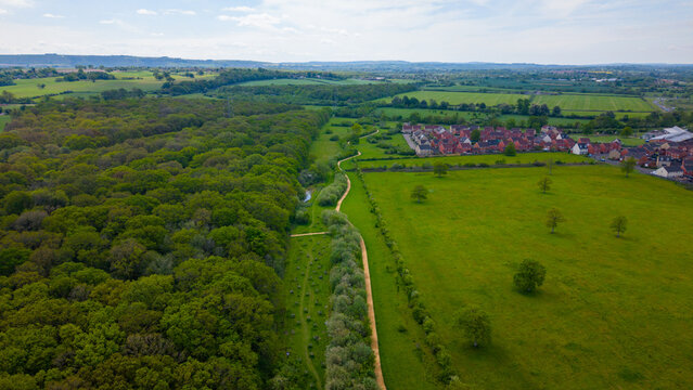 Aerial view of a winding path separating a dense forest from a lush green field with a residential housing development in the background in Melksham, Wiltshire, United Kingdom.