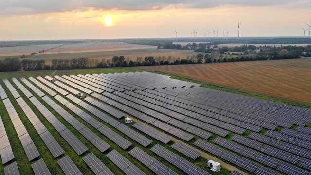 Aerial view of a large solar farm with rows of photovoltaic panels set in a rural landscape at sunset with wind turbines in the distance, Markisch-Oderland, Brandenburg, Germany.