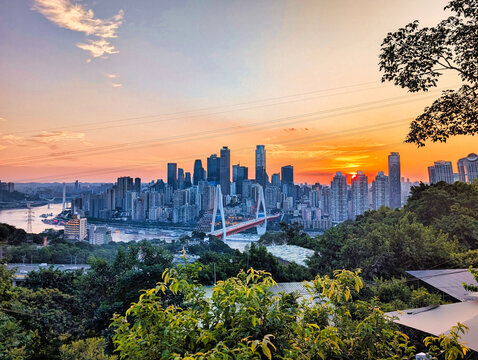View of the modern Chongqing skyline at sunset with the Caiyuanba Bridge spanning the Yangtze River, framed by lush hillside vegetation in Chongqing, Chongqing, China.