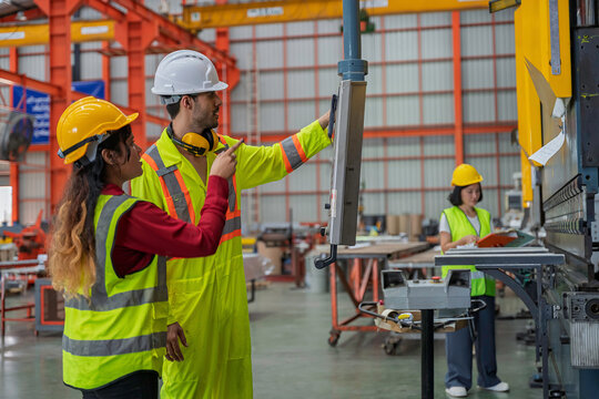 Technician and engineer review an industrial machine control panel while a worker operates equipment in a factory. Concept of technical training, teamwork, manufacturing, and production operations.