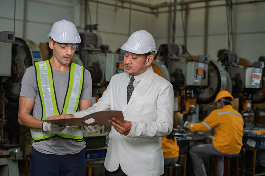 Senior Asian factory manager in a suit reviewing production documents with a male industrial engineer wearing safety gear on the manufacturing floor with other workers in the background.