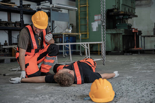 Panicked male industrial worker uses a walkie-talkie to urgently call for emergency help for an unconscious injured colleague lying on the ground inside a manufacturing factory.