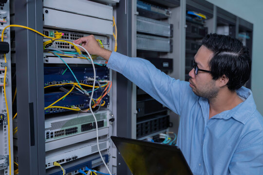 Focused Asian male IT network administrator uses a laptop to monitor server performance inside a bright data center managing complex corporate cybersecurity operations and infrastructure.