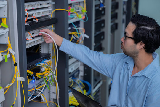 Focused Asian male IT network administrator uses a laptop to monitor server performance inside a bright data center managing complex corporate cybersecurity operations and infrastructure.