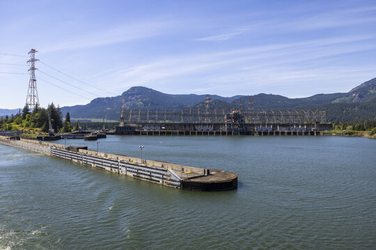 View of the Bonneville Dam on the Columbia River with concrete lock walls, power lines, and mountains in the background under a clear blue sky in Cascade Locks, Oregon, United States.
