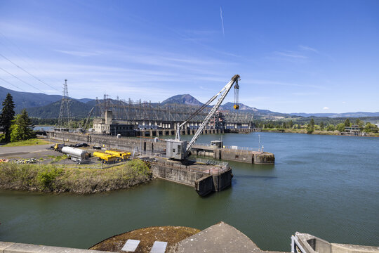 View of Bonneville Dam on the Columbia River with a large industrial crane, power lines, and mountains in the background under a clear blue sky, Cascade Locks, Oregon, United States.