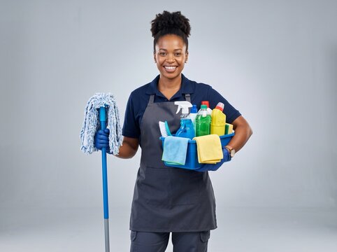 Smiling young woman cleaner holding a mop and a basket full of colorful cleaning supplies and chemical bottles for professional housekeeping services