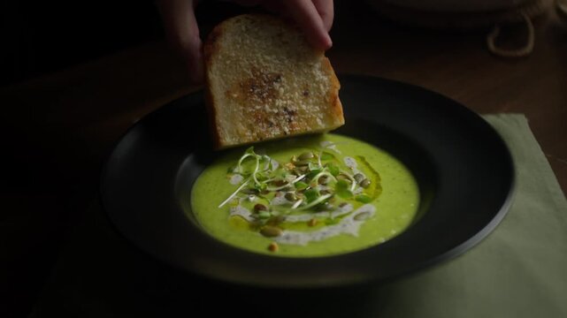 A chef dips crispy toasted bread into creamy zucchini soup garnished with microgreens, seeds, cream and olive oil, creating an elegant and appetizing meal.