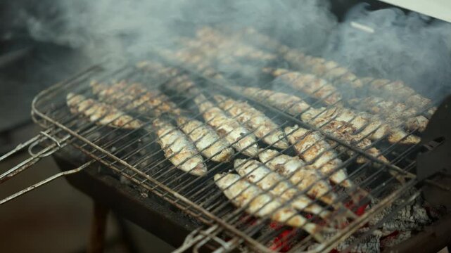 Small fish cooking on a charcoal barbecue grill, with smoke rising, preparing fresh seafood outdoors