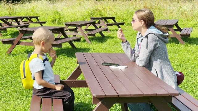 A mother and young boy sit at a wooden picnic table eating in a sunny park. They feel happy, joyful, and peaceful during this calm outdoor break in the green landscape.