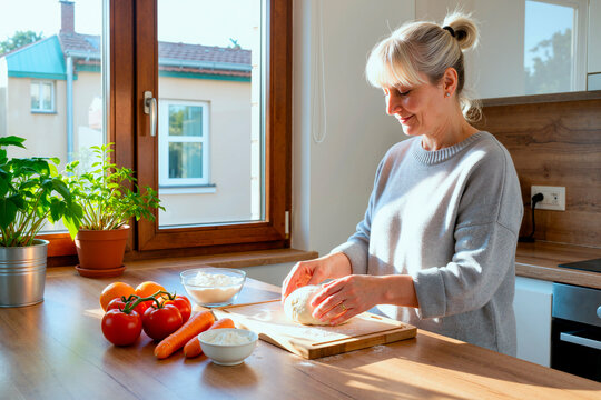 Caucasian middle aged woman kneading dough in home kitchen, preparing fresh meal with vegetables and flour. Supporting healthy cooking, home food content, lifestyle marketing