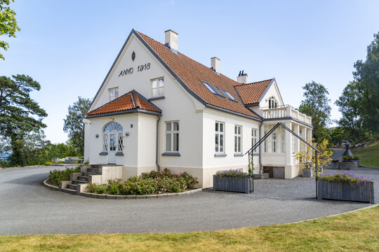 View of a historic white villa with a red tiled roof and the inscription Anno 1918 on the gable, set in a garden with a gravel driveway in Kristiansand, Agder, Norway.