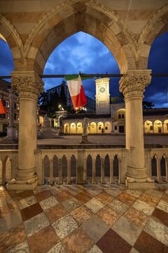Arcades of the Loggia del Lionello with Italian flags, illuminated main square Piazza della Libert&agrave; with Loggia e Tempietto di San Giovanni and clock tower, Castello di Udine behind, blue hour, dramatic sky, old town, Udine, Friuli-Venezia Giulia, Italy
