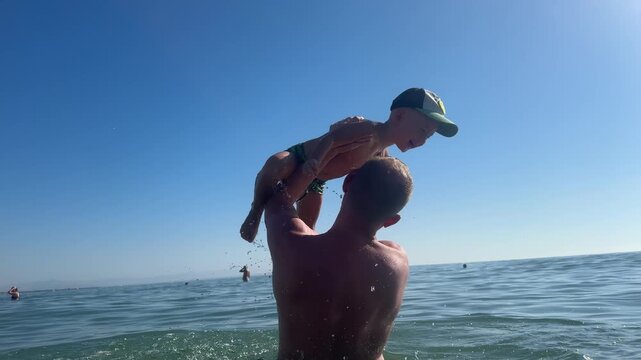 Father plays with his son in the sea, repeatedly lifting him out of the water and letting him jump back into the waves. 4k