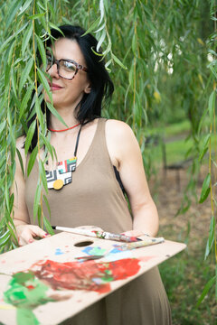 Young woman artist with artist palette by the willow tree in the forest, looking at the camera and smiling