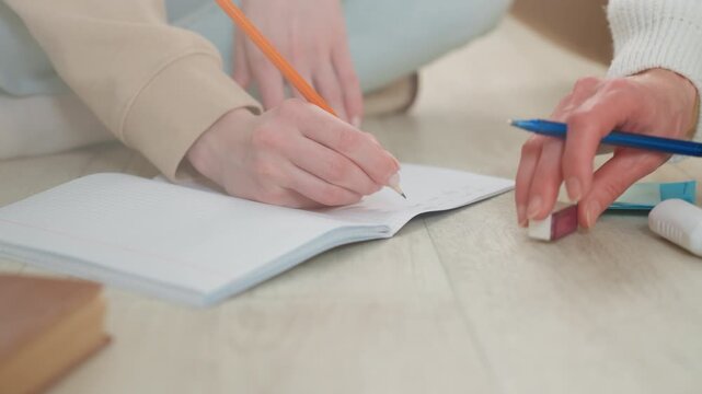 Female tutor writing grammar notes closeup of hands on wooden floor, pencil and eraser on open notebook, tutor guiding student through sentence exercises, warm daylight, casual sweater, calm
