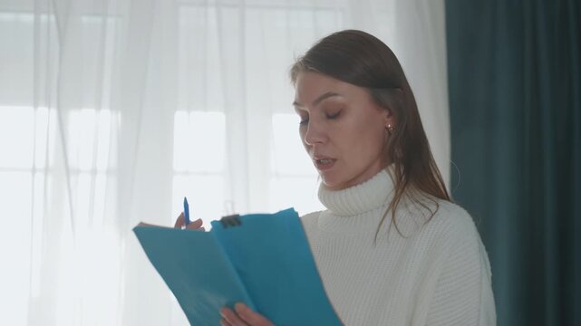 Woman rehearsing lines with blue binder, private coaching moment annotating script and correcting diction, thoughtful posture by curtain, soft daylight, intimate home setup for online lesson