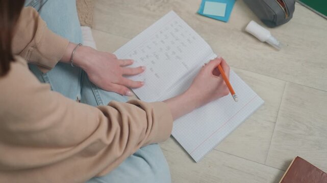 Learner completing grammar workbook on floor, steady pencil strokes, attentive notetaking, casual sweater, organized stationery pouch, calm concentration and steady progress during solo practice