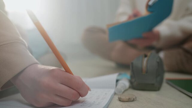 Close up pencil writing on notebook tutor holds blue notebook student sits crosslegged on floor pencil case and eraser nearby soft morning light gentle grammar coaching guided homework correction cozy