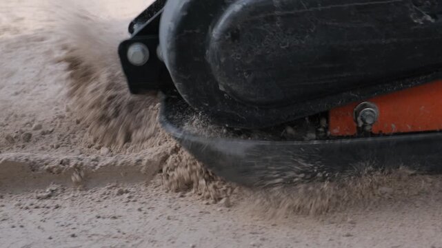 Close up of a vibratory plate compactor moving over a sand base, compacting it for sidewalk paving, vibrating sand bouncing and scattering to the sides, construction and urban improvement