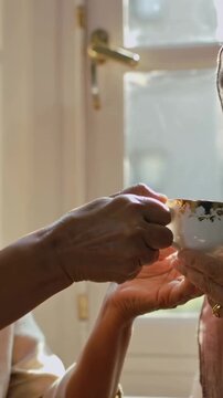 Vertical video: Offering left senior guiding teacup to right woman receiving on window seat for tea