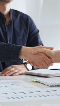 Business partners shaking hands after successful meeting in office while another person is clapping hands over financial documents, laptop and notebook