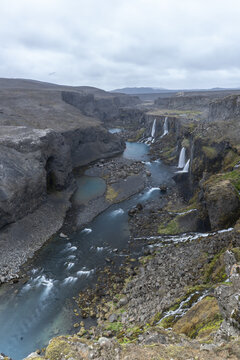 View of the Sigoldugljufur canyon with its multiple waterfalls cascading into a turquoise river surrounded by rugged volcanic rocks and mossy terrain in Iceland.