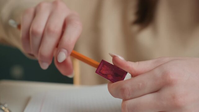 Closeup caucasian female sharpening pencil with handheld sharpener, shavings falling, focused fingers, preparing for homework, home study ritual, tactile detail, quiet morning light