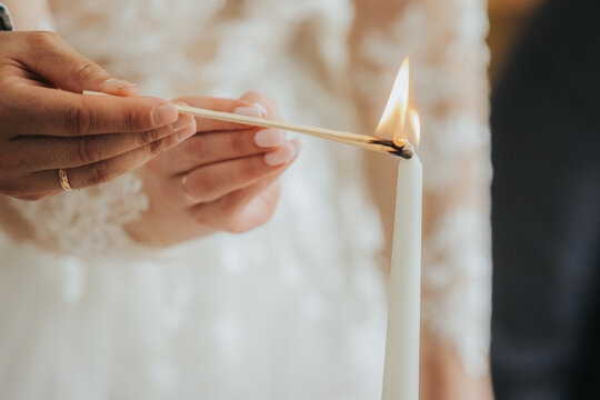 Bride lighting a white candle with a match during a wedding ceremony, delicate lace details on her dress and soft lighting creating an intimate atmosphere