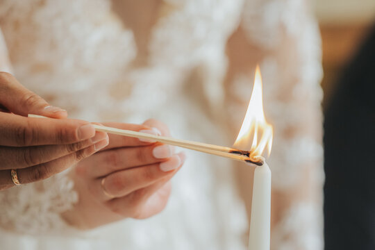 Bride lighting a white candle with a matchstick during a wedding ceremony, wearing a lace dress, with soft lighting creating a warm atmosphere in the background
