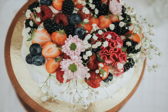 Colorful cake topped with fresh strawberries, blueberries, blackberries, and decorative flowers on a golden cake board, displayed on a wooden table