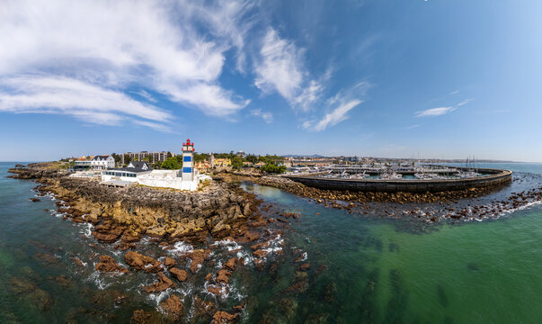 Aerial view of Farol Museu de Santa Marta lighthouse on the rocky coastline next to the marina with boats in the harbor under a clear blue sky in Cascais, Lisboa, Portugal.