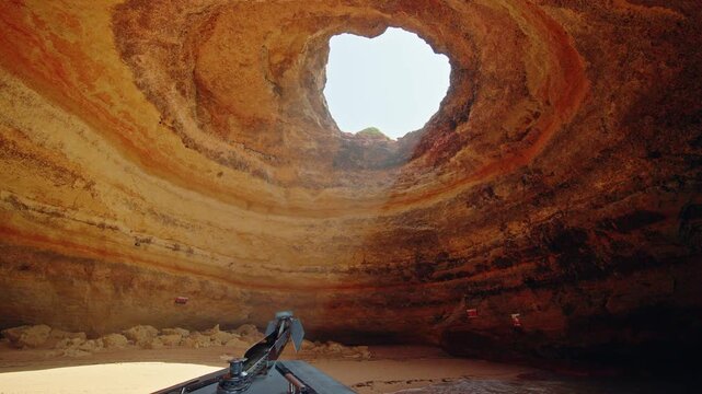 View from a tour boat inside the famous Benagil Cave with a sandy beach, layered limestone walls and a natural ceiling opening.
