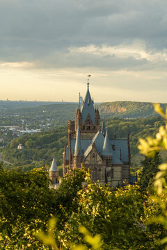 View of Schloss Drachenburg with its pointed towers and stone architecture surrounded by lush forests and rolling hills under a soft sunset sky in Konigswinter, North Rhine-Westphalia, Germany.