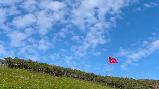 Large red Turkish flag waving in the wind above the forested hills of the Princes Islands in Istanbul, bright blue sky with scattered white clouds creating open airy atmosphere