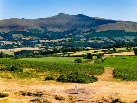 Elevated view of Pen y Fan peak from Mynydd Illtud, triangulation station in foreground overlooking rural Welsh valleys and rolling hills beneath clear blue sky
