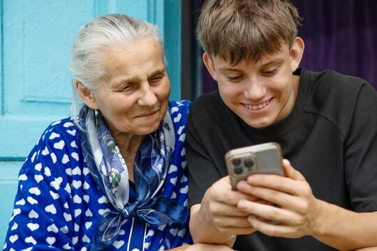 Happy teenage grandson and senior grandmother looking at mobile phone screen, smiling and sharing warm family moment outside.