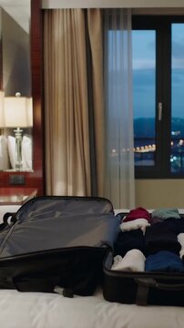 Vertical video: Panning camera showing open black suitcase on bed in hotel room with packed clothes