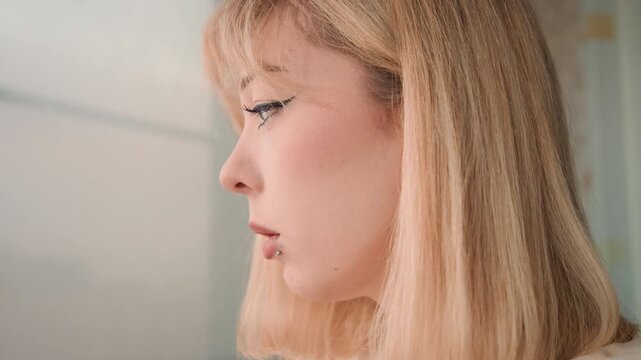 Female indoors smoking. Woman smokes thoughtfully inside. Lady with lip piercing smokes while sitting near window. Woman with lip piercing exhales smoke thoughtfully near window inside