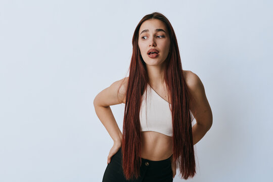 Young woman with long braided hair wearing white crop top and black pants posing with hand on back and looking sideways on plain background for fashion or lifestyle concept.