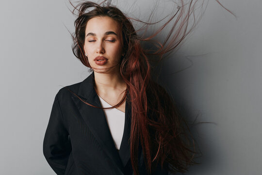 Woman with long hair blowing in the wind wearing black blazer and white shirt against gray background. Portrait with closed eyes and natural makeup for fashion or beauty concept.