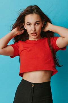 Young woman posing with hands in hair wearing red crop top and black pants against blue background. Casual style, confident expression and long dark hair with natural look.