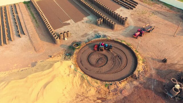 Aerial view of a tractor mixing clay in a circular pit at a brick manufacturing site with stacks of bricks and grain drying in the sun in Vijayawada, Andhra Pradesh, India.