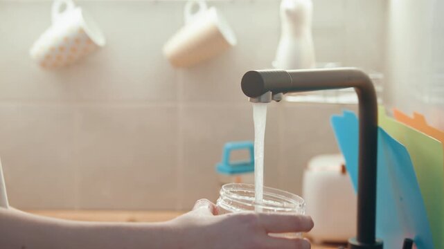 Hands rinsing jar under running faucet in bright kitchen weekend baker clears residue from glass container, steady stream and colorful boards create calm ingredient preparation scene